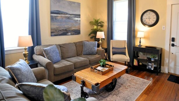 A living room with a sofa set, a coffee table, a side table with a lamp, a chair, a painting on the wall, and a clock. - The Hill District House - Kid Friendly, Walkable - Stay Smiley Rentals (St. Louis)