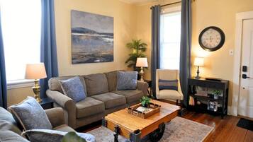 A living room with a sofa set, a coffee table, a side table with a lamp, a chair, a painting on the wall, and a clock.