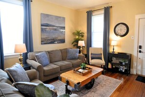 A living room with a sofa set, a coffee table, a side table with a lamp, a chair, a painting on the wall, and a clock.