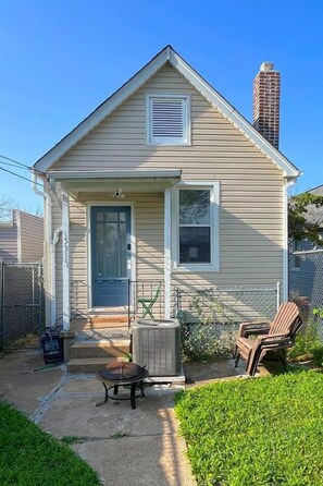 A single-story house with a front porch, a small outdoor seating area, and a tall building in the background.