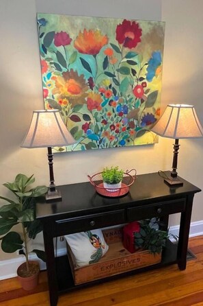 A black console table with two lamps, a floral painting, and a potted plant.