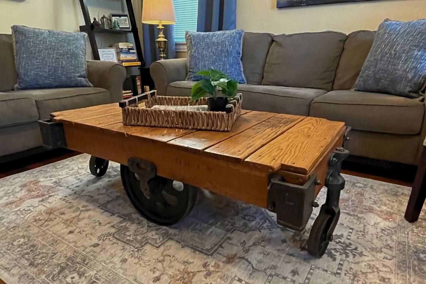 A wooden cart table with wheels, a plant, and a basket in a living room with a patterned rug, a sofa, and a lamp.