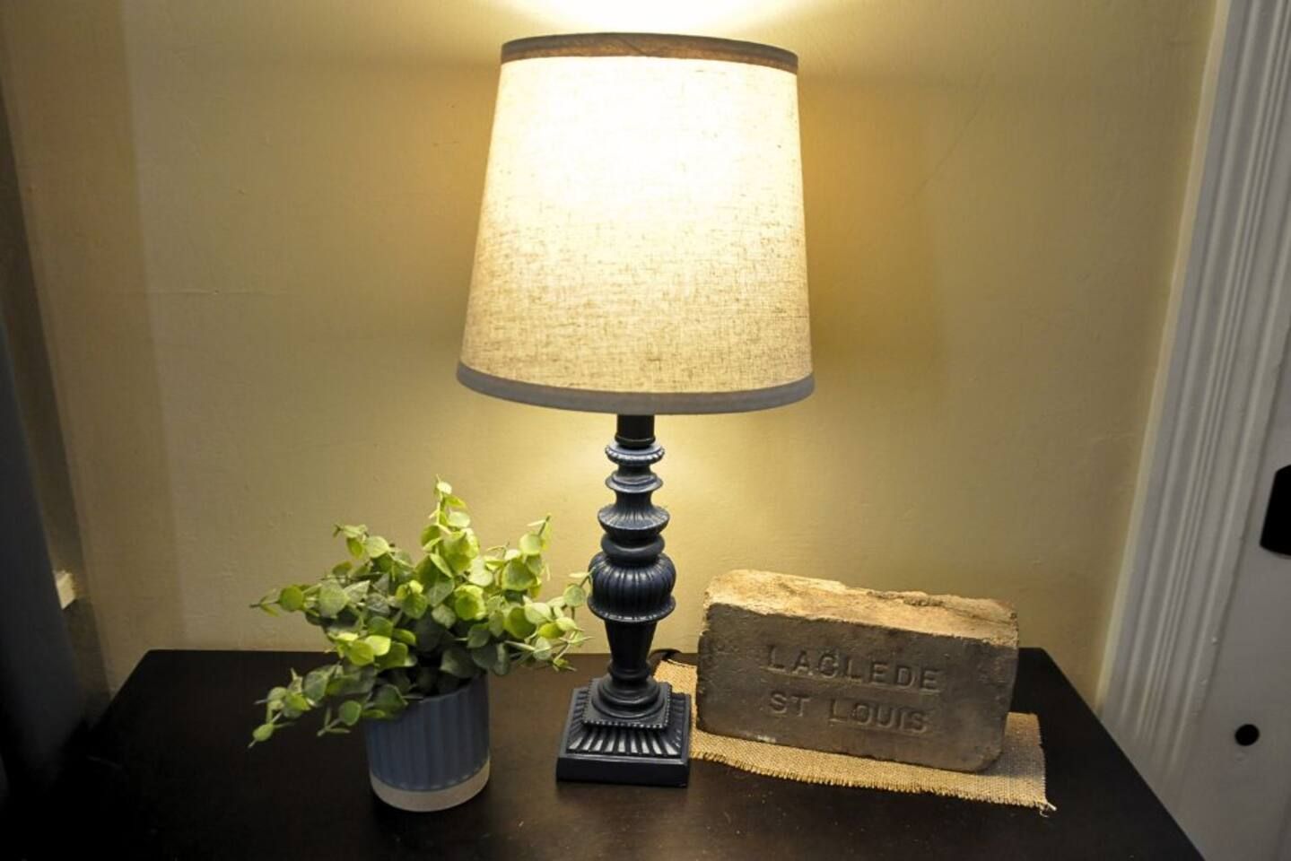 Een tafel met een lamp, een potplant en een steenblok waarop 'LAGUDE ST. LOUIS' gegraveerd staat.