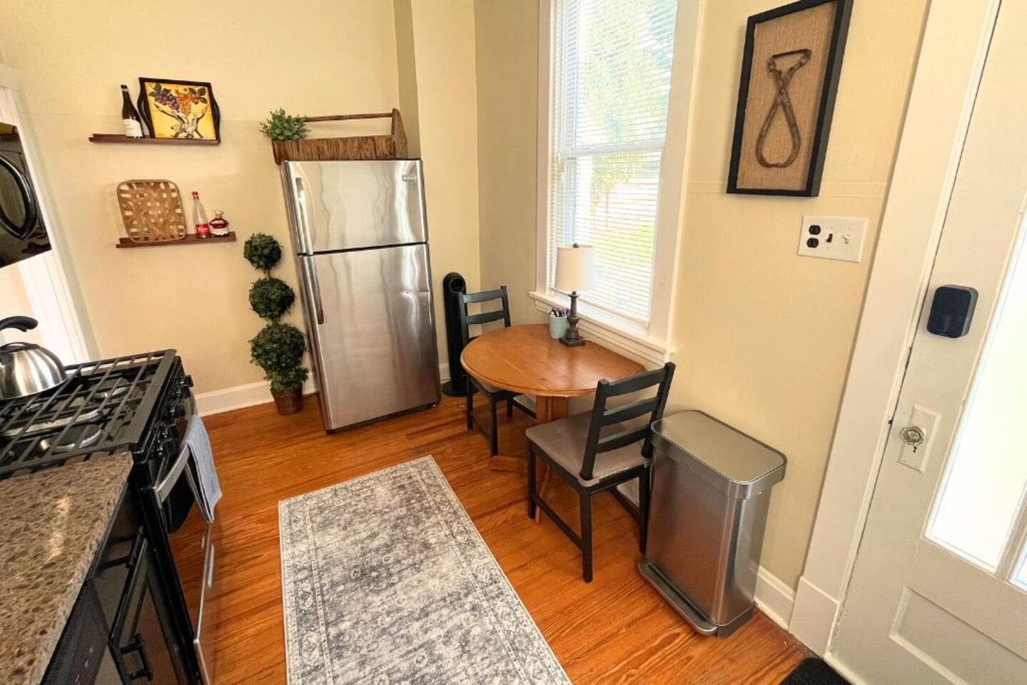 A kitchen with a stainless steel refrigerator, wooden dining table, chairs, a rug, and a window with blinds.