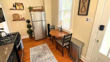 A kitchen with a stainless steel refrigerator, wooden dining table, chairs, a rug, and a window with blinds.