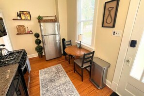 A kitchen with a stainless steel refrigerator, wooden dining table, chairs, a rug, and a window with blinds.