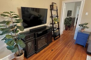 A living room with a flat-screen TV on a wooden cabinet, a ladder shelf with decorative items, and a potted plant.
