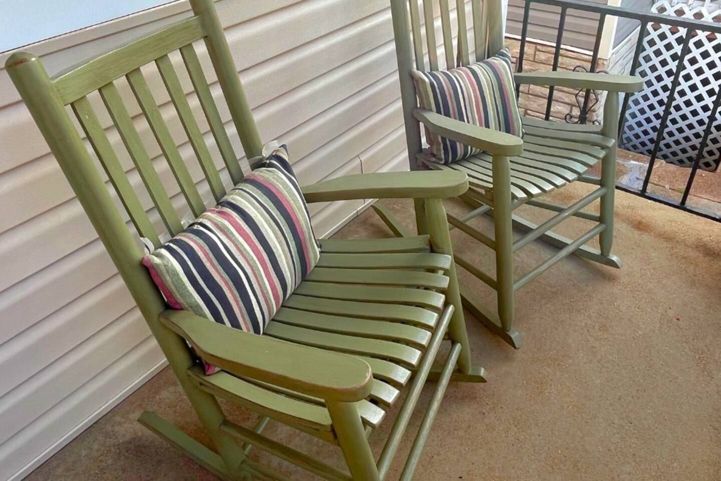 Deux fauteuils à bascule verts, dotés de coussins à rayures, se trouvent sur un balcon orné d’une rambarde en métal.
