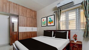 A bedroom with a bed, wooden wardrobe, two framed artworks, a window with curtains, and a small bedside table with a vase. - Hotel Central Vishwanathapuram Formerly (Coimbatore)