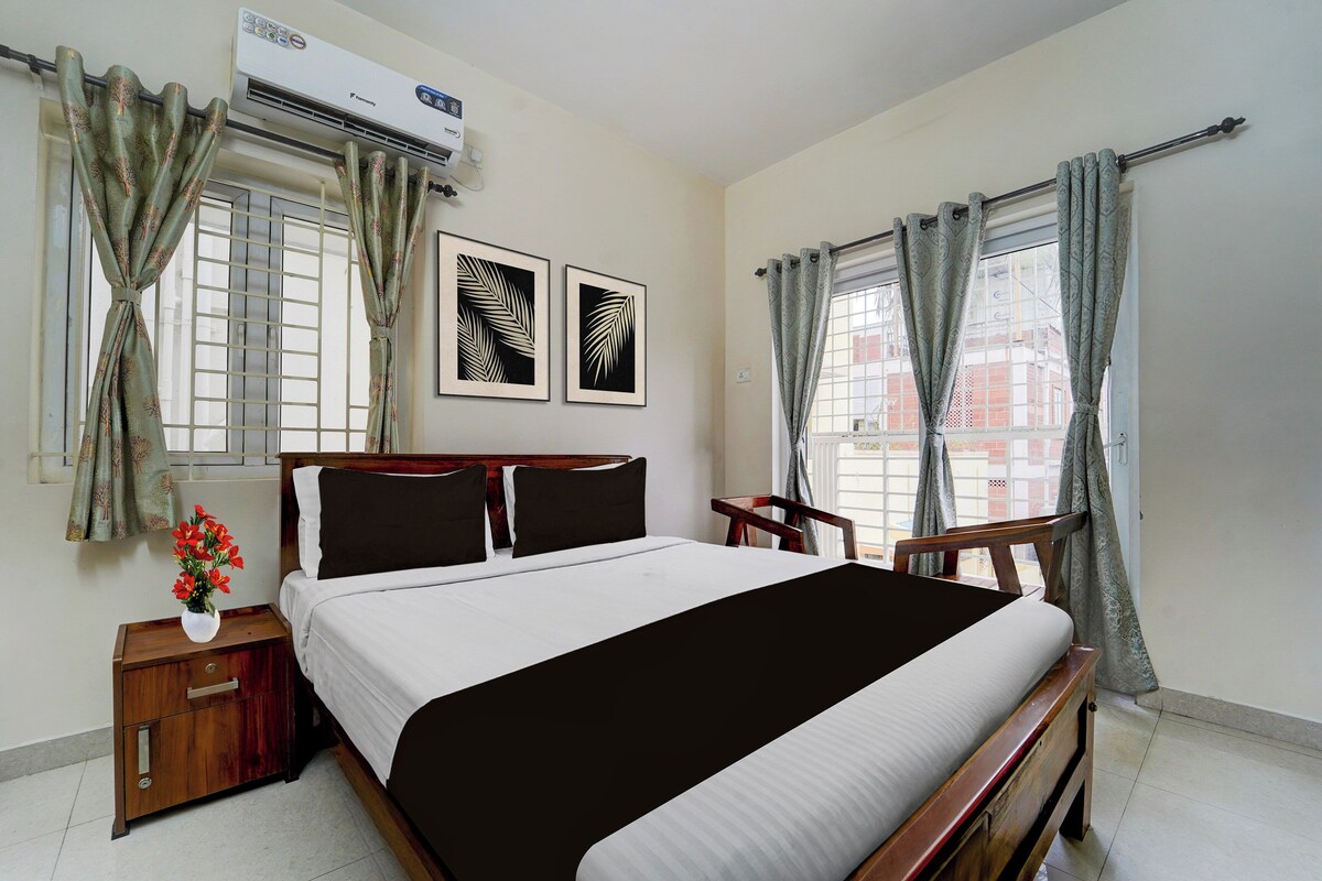 A hotel room with a bed, a nightstand, a chair, a window with curtains, and a wall-mounted air conditioner.