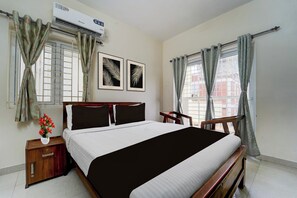 A hotel room with a bed, a nightstand, a chair, a window with curtains, and a wall-mounted air conditioner. - Hotel Central Vishwanathapuram Formerly (Coimbatore)