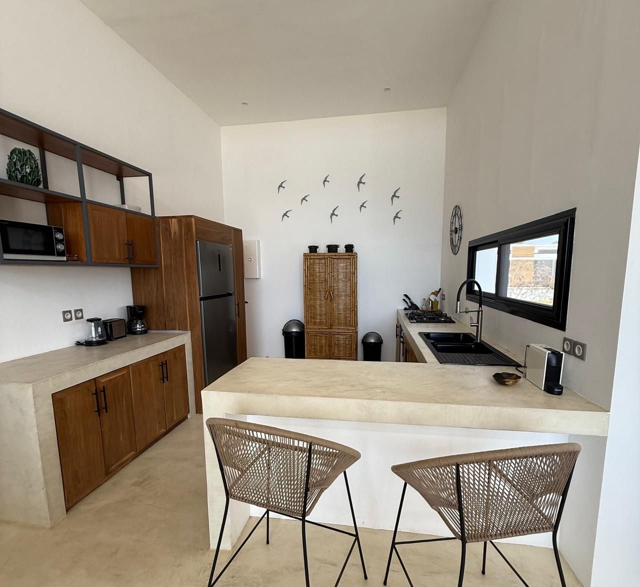 Une cuisine moderne avec des meubles en bois, un îlot central, deux chaises en osier et une horloge murale.
