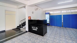 Ein Hotel-Empfangsbereich mit einem schwarzen Empfangstresen, einer Uhr, einem Ventilator und blauen Schließfächern im Hintergrund.
