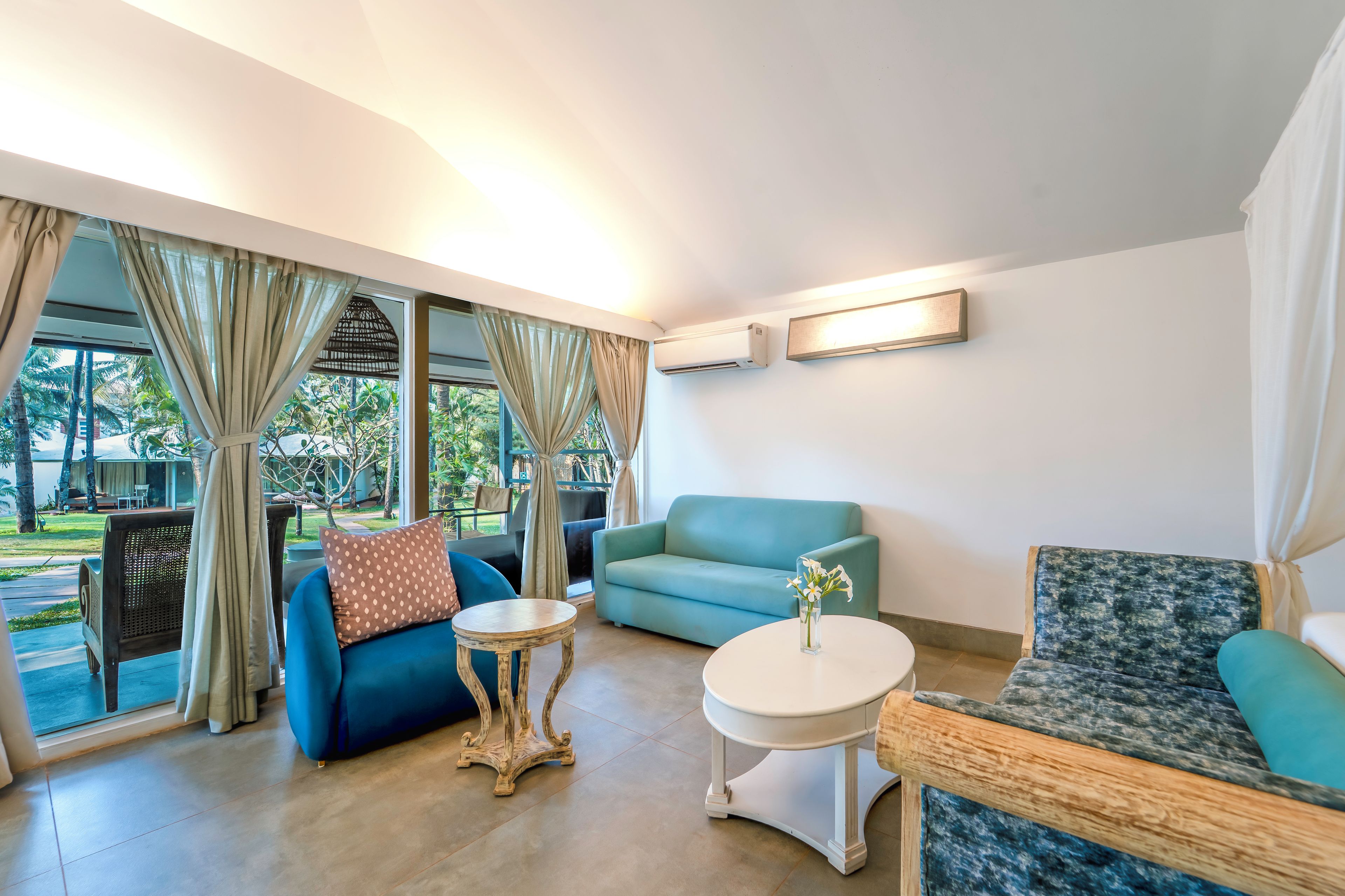 Una habitación con un sofá azul, una mesa redonda y una silla estampada. Se ve una ventana con cortinas y vistas a vegetación desde el exterior.