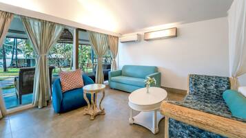 Una habitación con un sofá azul, una mesa redonda y una silla estampada. Se ve una ventana con cortinas y vistas a vegetación desde el exterior.