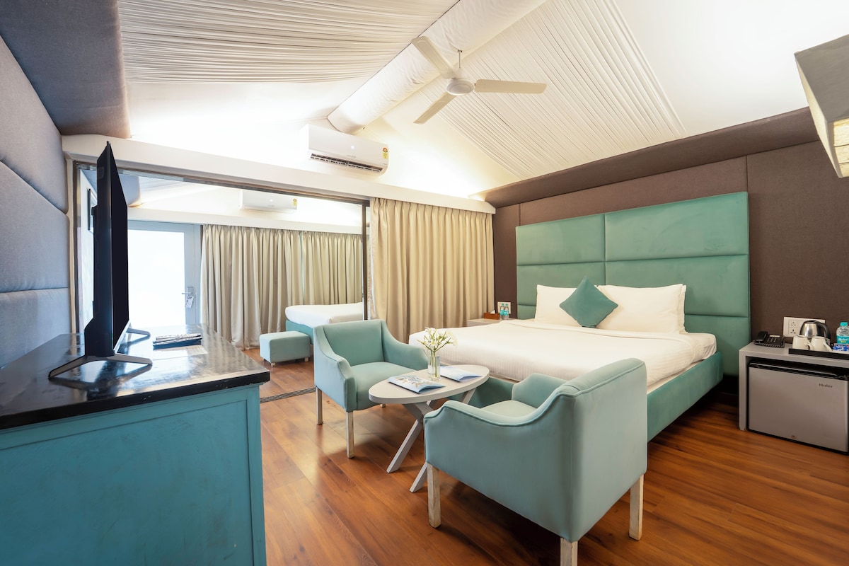 A hotel room with a bed, a desk, a chair, a small table, a minibar, and a ceiling fan.