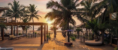 A poolside area with lounge chairs and a wooden deck, overlooking a beach with palm trees.