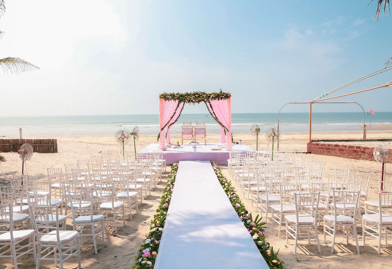 Une cérémonie de mariage organisée sur la plage, avec une allée blanche, des chaises et une scène décorée de draperies roses.