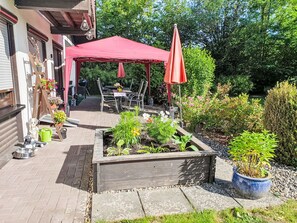 Een terras met een rode parasol, zitplaatsen buiten en een tuin met verhoogde bloembedden.