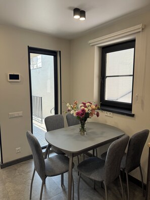 Uma sala de jantar com uma mesa redonda, quatro cadeiras cinzas, um vaso com flores e uma janela com persianas.