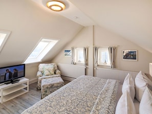 Ein Schlafzimmer mit schrägverdeckter Decke, einem Bett mit gemusterten Textilien, einem Fernseher auf einem Schrank, einem Sessel und zwei Fenstern mit Vorhängen.