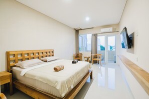 Quarto de hotel com cama de madeira, uma pequena mesa, uma cadeira, televisão e porta de vidro deslizante para uma varanda.