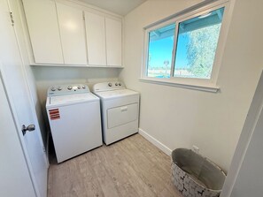 A laundry room with two white washers and dryers, a window with a view of greenery, and a wooden floor. - 8651-Peaceful 4-bedroom house with AC in charming Elk Grove (Elk Grove)