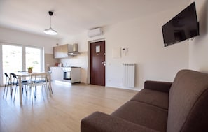 Ein Wohnzimmer mit braunem Sofa, eine Essecke mit Tisch und Stühlen, eine Küche mit Schränken und Herd sowie ein an der Wand befestigter Fernseher.