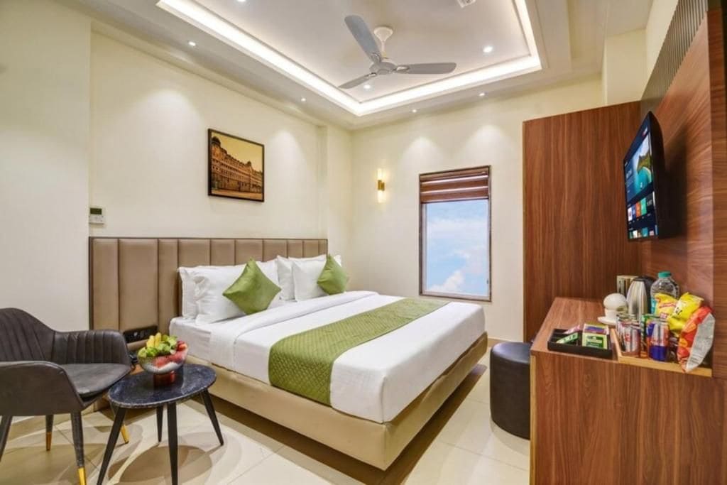 A hotel room with a bed, a desk, a chair, a small table with a fruit bowl, a ceiling fan, a wall-mounted TV, and a window with a view.