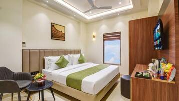 A hotel room with a bed, a desk, a chair, a small table with a fruit bowl, a ceiling fan, a wall-mounted TV, and a window with a view.
