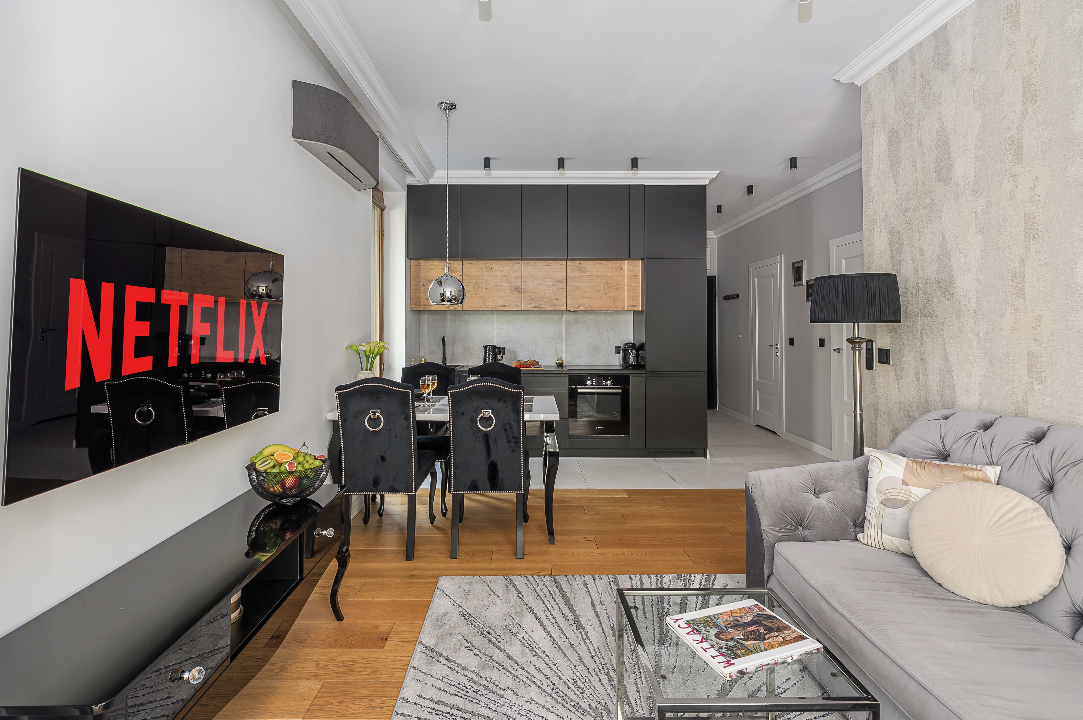 Un salon moderne doté d’un téléviseur à écran plat, d’un coin repas avec des chaises, d’une cuisine avec un plan de travail en bois et d’un canapé gris sur lequel repose un magazine sur une table basse.