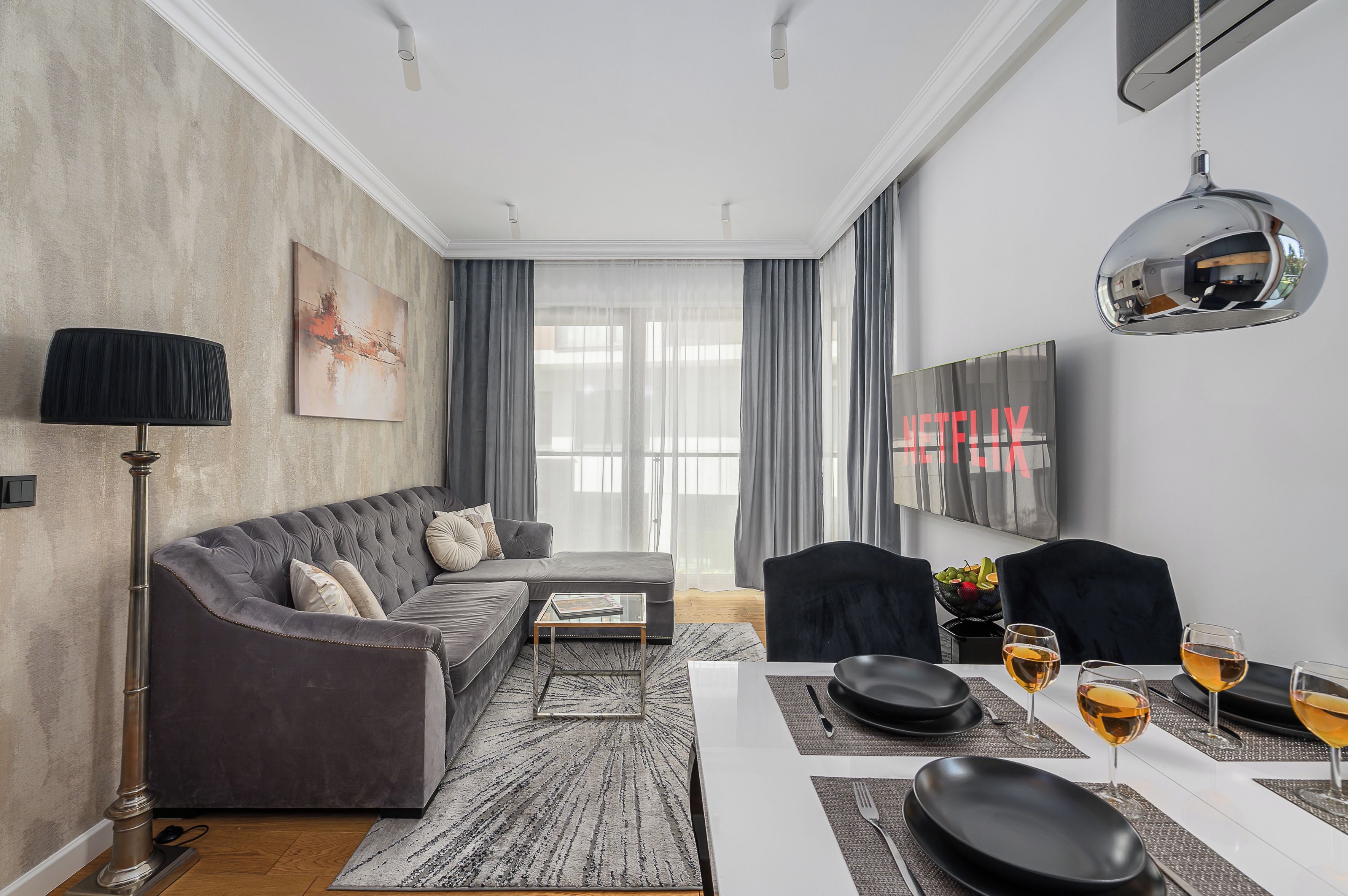A modern living room with a grey sofa, a coffee table, a dining table set for two, and a television mounted on the wall.