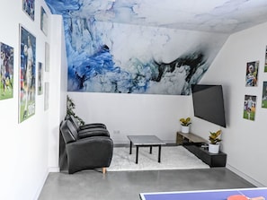 Una sala de estar moderna con un gran mural abstracto, un televisor de pantalla plana, un sofá de cuero negro, una mesita de centro pequeña y una mesa de ping pong.