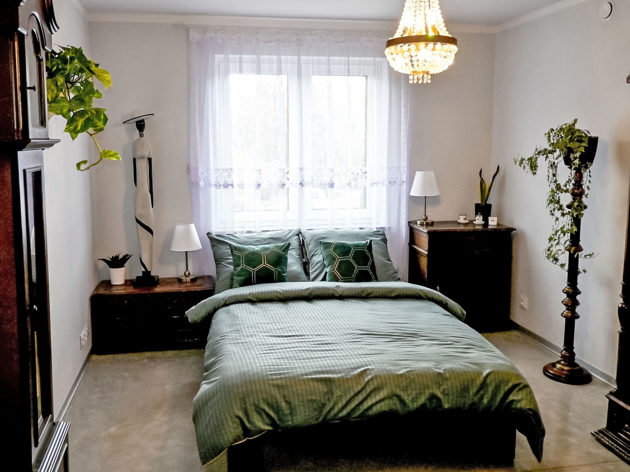 Quarto com cama, mesas de cabeceira, luminária, janela com cortinas e planta pendente.