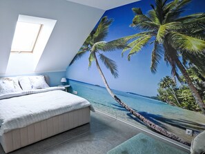 A bedroom with a bed, a nightstand, a lamp, a window, and a wall mural of a beach scene.