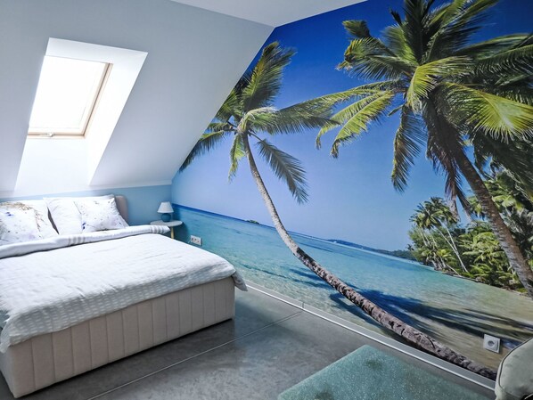 A bedroom with a bed, a nightstand, a lamp, a window, and a wall mural of a beach scene.