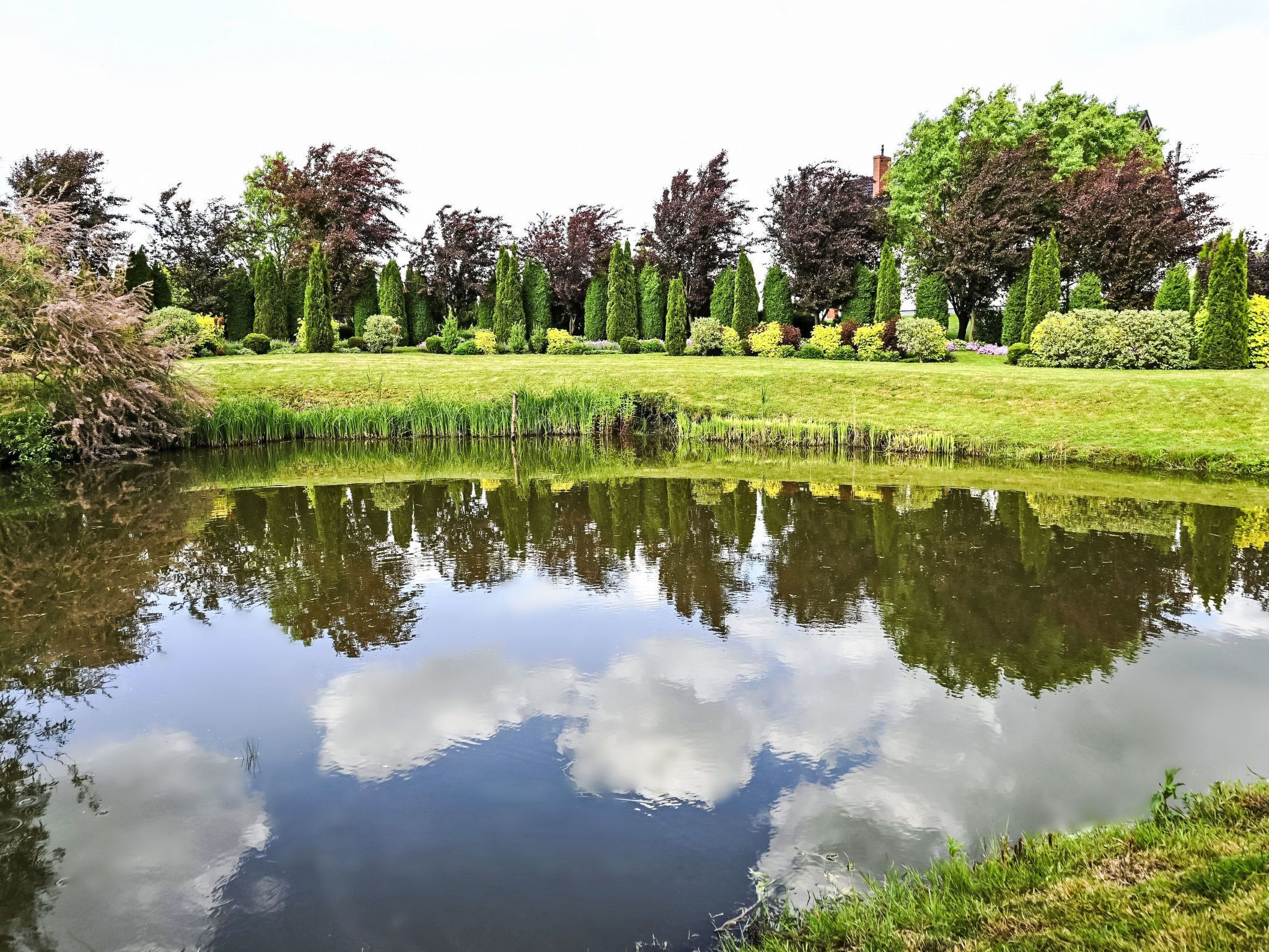 Un étang paisible dont les eaux reflètent clairement les arbres et le ciel, entouré de pelouses et de haies bien entretenues.