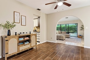 Ein modernes Interieur mit einem Holzkonsolentisch, gerahmten botanischen Drucken, einem Deckenventilator und Blick in einen Wohnbereich mit einer Couch und großen Fenstern.