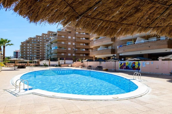 A swimming pool with a thatched roof overhead, surrounded by multi-story buildings with balconies. - Oropesa Apartment with Terrace (Orpesa)