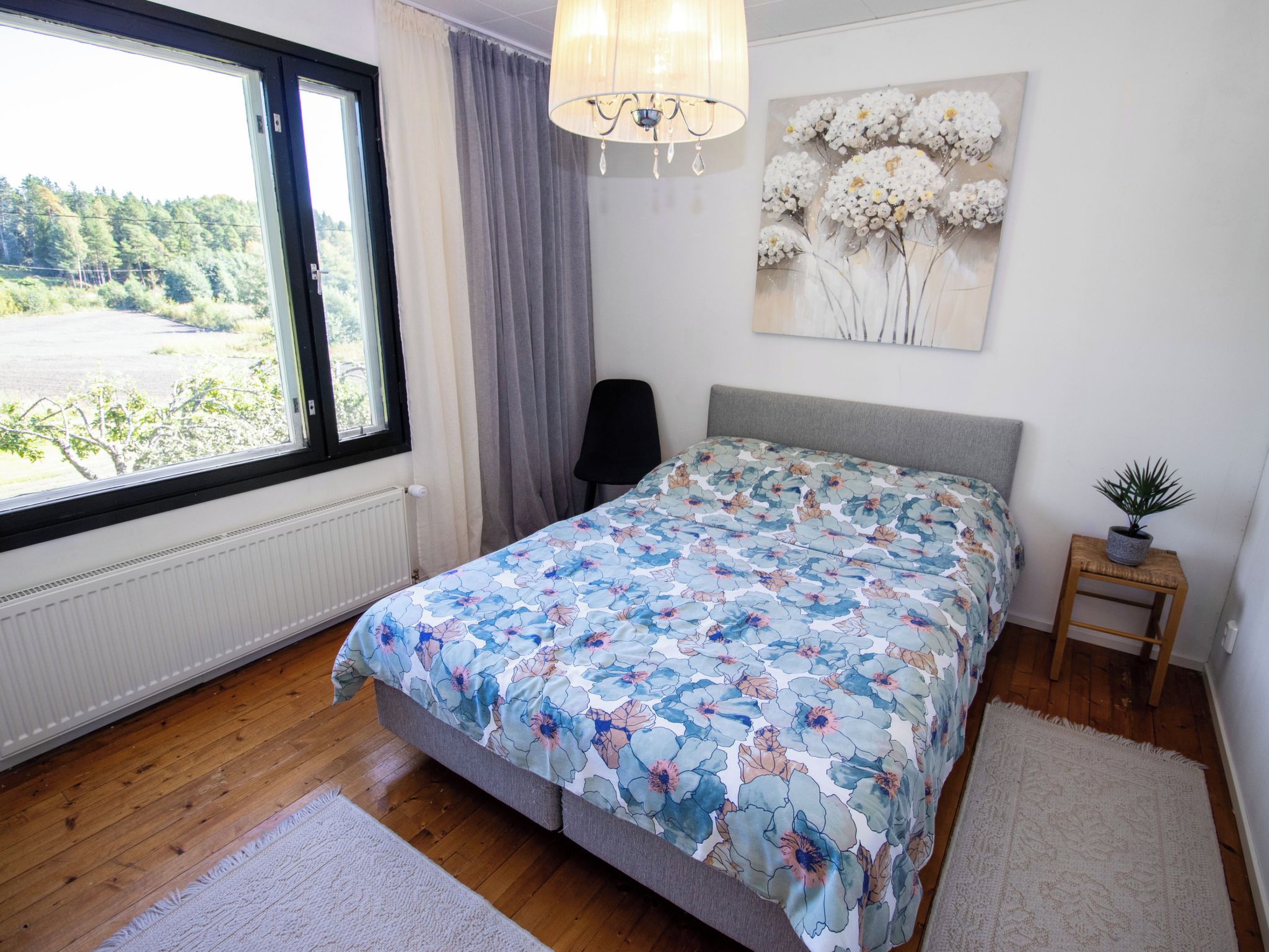 Une chambre avec une grande fenêtre, un lit recouvert d’une literie à motifs floraux, une chaise, une petite table avec une plante et une œuvre d’art murale.