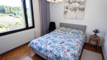 Une chambre avec une grande fenêtre, un lit recouvert d’une literie à motifs floraux, une chaise, une petite table avec une plante et une œuvre d’art murale.