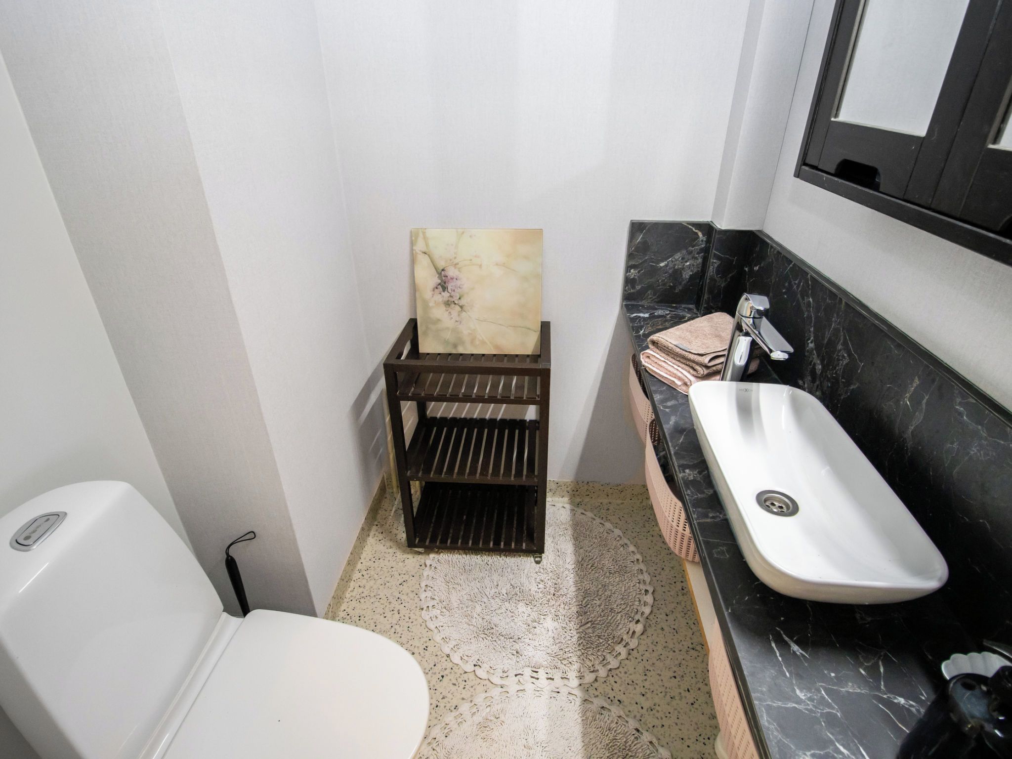 Ein kleines Badezimmer mit einer weißen Toilette, einem schwarzen Marmorwaschbecken und einem Holzregal mit einem Gemälde.