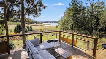 Eine Holzterrasse mit einer Couch und einem Tisch, mit Blick auf eine natürliche Landschaft mit Bäumen und einem Gewässer.