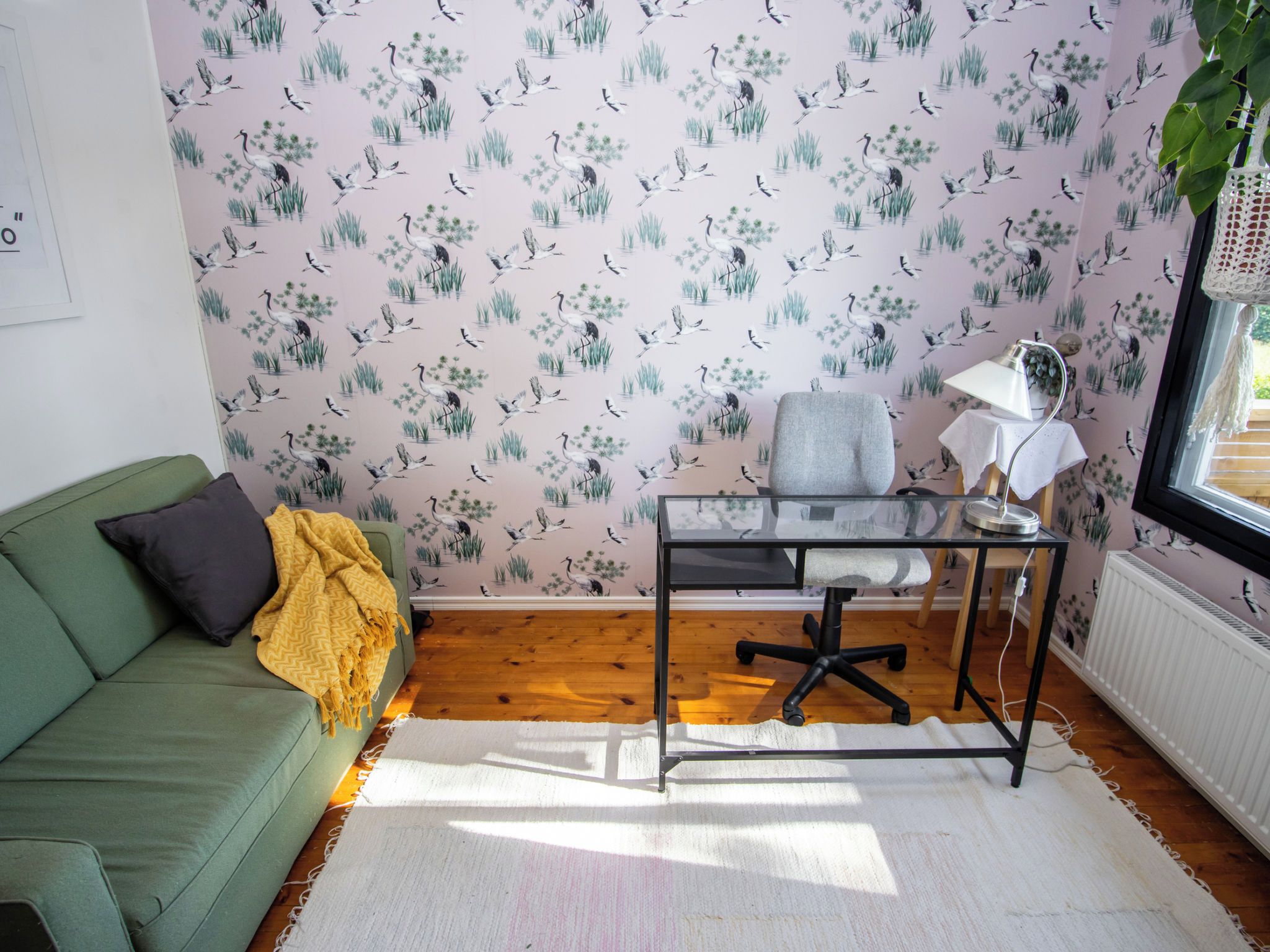 Une pièce avec un canapé vert, une petite table avec une lampe et un mur recouvert d’un papier peint à motifs d’oiseaux.