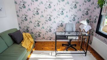 Une pièce avec un canapé vert, une petite table avec une lampe et un mur recouvert d’un papier peint à motifs d’oiseaux.