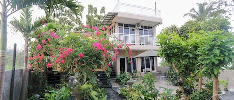 A two-story building with a balcony, surrounded by lush greenery and flowering plants.