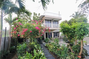 A two-story building with a balcony, surrounded by lush greenery and flowering plants.