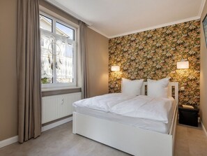 A bedroom with a bed, a window with curtains, a radiator, and a floral wallpaper. - 8, Residence Dunenstrasse (Binz)