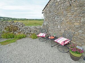 An outdoor seating area with stone walls, two metal chairs with striped cushions, a table with a bottle and glass, and a flower pot with pink flowers. - 1 bedroom accommodation in Carr Bank, Arnside (Carr Bank, Arnside)