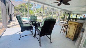 Een overdekte patio met een tafel, stoelen, een plafondventilator en uitzicht op een zwembad.